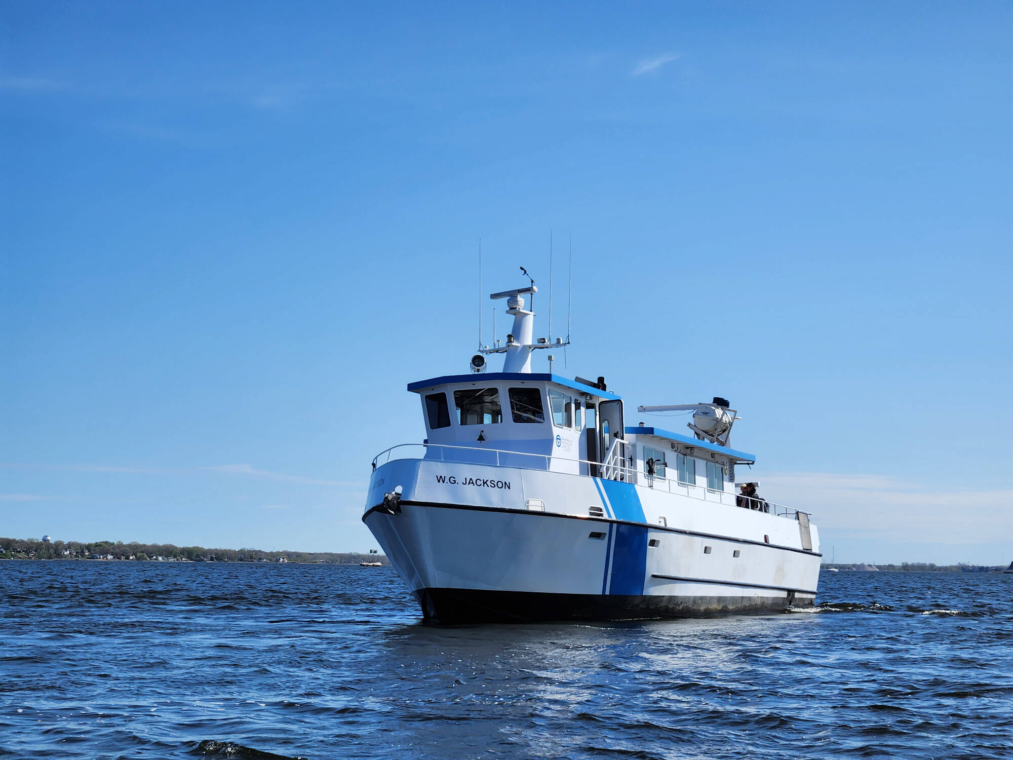 The W.G. Jackson research vessel sails on Muskegon Lake. The boat is white with a blue roof and blue stripe on the side. The sky and water are bright blue.